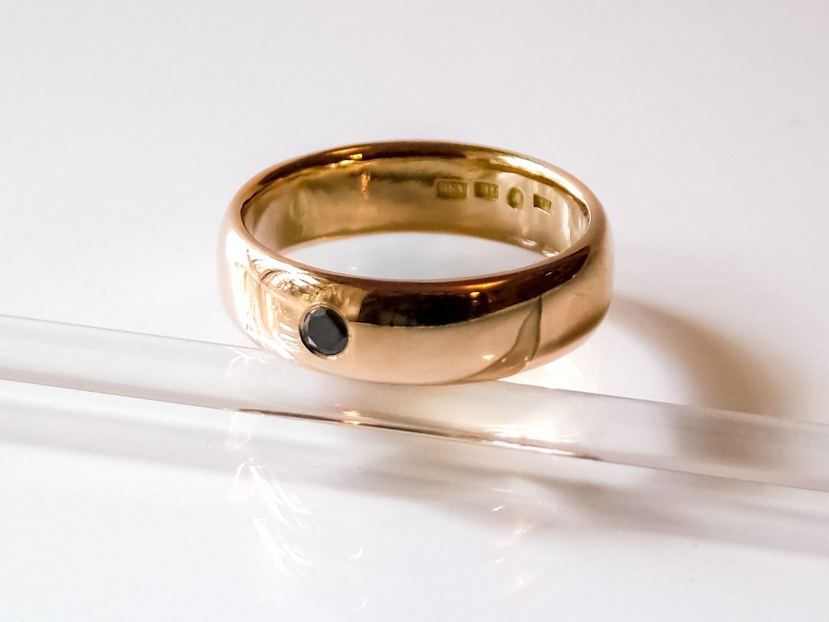 Sample photo used in the jewelry identification demo: a wide domed band in warm yellow metal with one small dark round stone set flush in its outer face, resting on a clear acrylic rod against a white background, with a row of small punch marks catching the light inside the band
